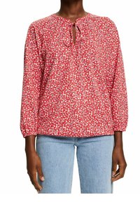 Blusa rossa floreale con scollatura tonda, dettaglio a fiocco e maniche lunghe elastiche. Il tessuto presenta piccoli fiori bianchi e blu su uno sfondo vivace.