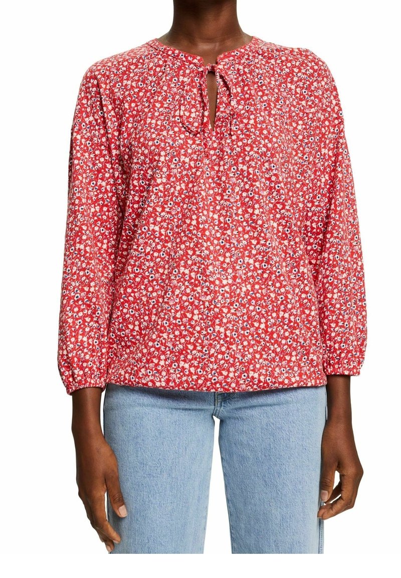 Blusa rossa floreale con scollatura tonda, dettaglio a fiocco e maniche lunghe elastiche. Il tessuto presenta piccoli fiori bianchi e blu su uno sfondo vivace.