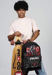 La mochila para patineta cuenta con tela negra y gráficos en rojo y blanco. La patineta de madera tiene un acabado suave con grandes ruedas naranjas y un logotipo.