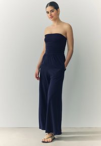 Mørkeblå stramfri jumpsuit med rynket bærestykke, elastisk talje, vide bukser og lommer. Båret med flade sandaler med metallic detaljer.