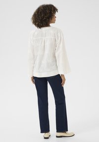 Blusa bianca ricamata con maniche lunghe e una vestibilità rilassata, abbinata a jeans blu scuro e calzature di colore chiaro.
