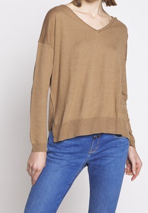 Kvinde iført en lysebrun V-hals trøje med lange ærmer, kombineret med blå denimjeans, vist fra nakke til hofter.