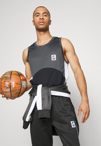 Muška sportska odjeća Nike sastoji se od sive majice bez rukava s bijelim i crnim uzorkom, uparenih s tamnosivim trenirkama i narančastom košarkaškom loptom.
