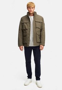 Olivengrøn isoleret jakke med høj krave, fire forlommer, let design, og kombineret med en lys grå sweater og marineblå bukser.