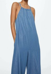 Salopette en jean à fines bretelles, avec un col froncé et des poches latérales. Présente une coupe décontractée et une couleur bleu clair avec une texture lisse.