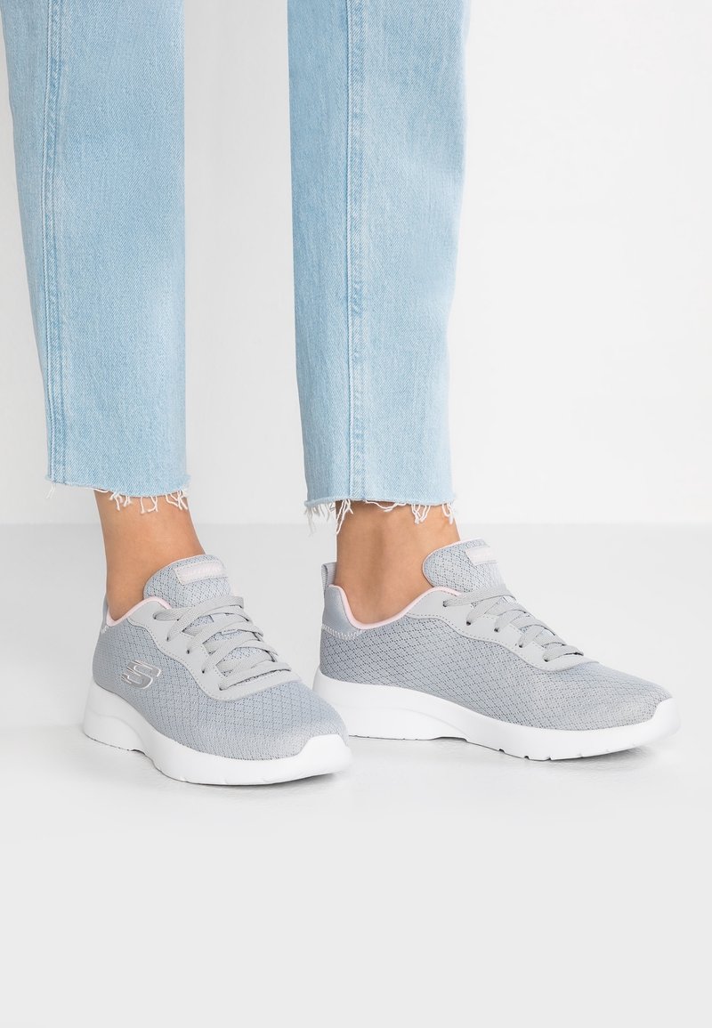Pieds portant des baskets bleu clair avec des accents roses et des semelles blanches, associées à un jean bleu clair à ourlet effiloché, sur fond blanc.