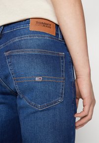 Nahaufnahme einer Person, die blaue Tommy Jeans Denim trägt, mit Logo-Patch am Bund und kleinem Fahnenemblem auf der Gesäßtasche.