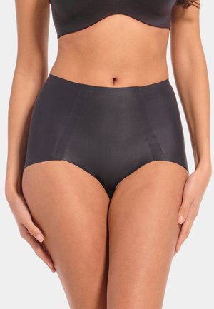 Femme portant des sous-vêtements noirs sans couture taille haute et un soutien-gorge assorti, debout les mains détendues le long du corps sur un fond uni.