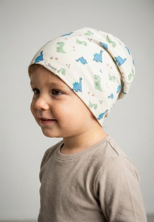Toddler wearing a cream slouchy hat patterned with blue and green dinosaurs and a beige long-sleeve shirt, smiling and looking slightly left.