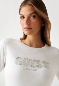 Una donna con capelli castano chiaro indossa un maglione bianco con la scritta "GUESS EST. 1981" decorata con strass sul petto.