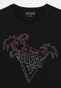 Camiseta de algodón orgánico negra con un diseño triangular que presenta el texto "GUESS" y gráficos de palmeras adornados con rhinestones rosas y blancos.