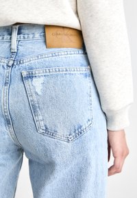 Lichtblauwe denim jeans met lichte beschadigingen op de achterzak; heeft een tanleren Calvin Klein Jeans logo patch op de tailleband.