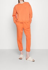 Felpa arancione e pantaloni della tuta abbinati realizzati in tessuto morbido, con scollo rotondo e polsini a coste. Abbinati a sandali beige pelosi a fascia.