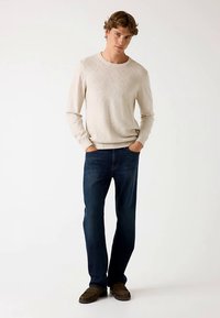 Svetlobéžový pletený sveter s textúrovaným diamantovým vzorom, kombinovaný s tmavomodrými džínsami a hnedými mokasínovými topánkami.