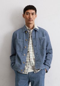 Leren spijkerjack in lichtblauw, met twee borstzakken en tan kleurige knopen, over een wit en geruit shirt, gecombineerd met bijpassende broek.