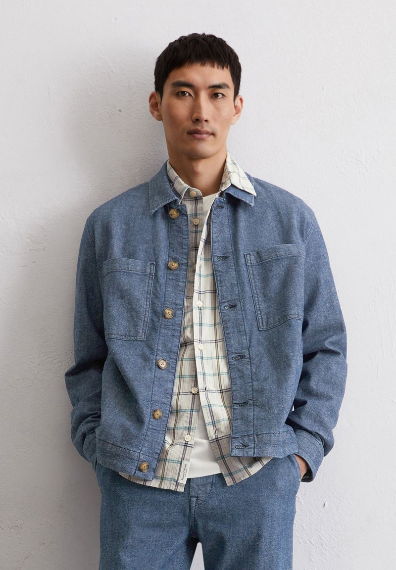 Leren spijkerjack in lichtblauw, met twee borstzakken en tan kleurige knopen, over een wit en geruit shirt, gecombineerd met bijpassende broek.