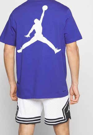 Mann trägt ein lila Shirt mit weißer Silhouette eines springenden Basketballspielers und weiße Basketballshorts mit schwarzen Streifen.