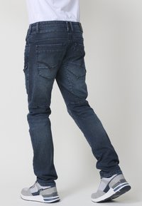Jeans de mezclilla de color azul oscuro con un corte ajustado, que cuentan con cinco bolsillos, costuras visibles y detalles de costura sutiles. Llevados con zapatillas deportivas grises.