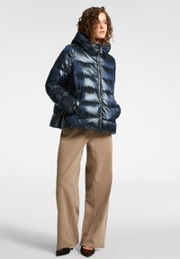 Chaqueta de plumón azul marino con textura brillante y acolchada, cierre de cremallera y capucha. Combinada con pantalones de pierna ancha beige, con un acabado de costura uniforme.