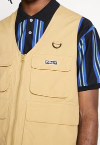 Beige zip-up väst med flera framfickor och en ringdetalj. Bärs över en blå, svart och grönrandig skjorta med krage.