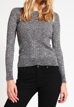 Femme portant un pull gris côtelé ajusté à manches longues et un pantalon noir taille haute, debout les mains relâchées le long du corps.