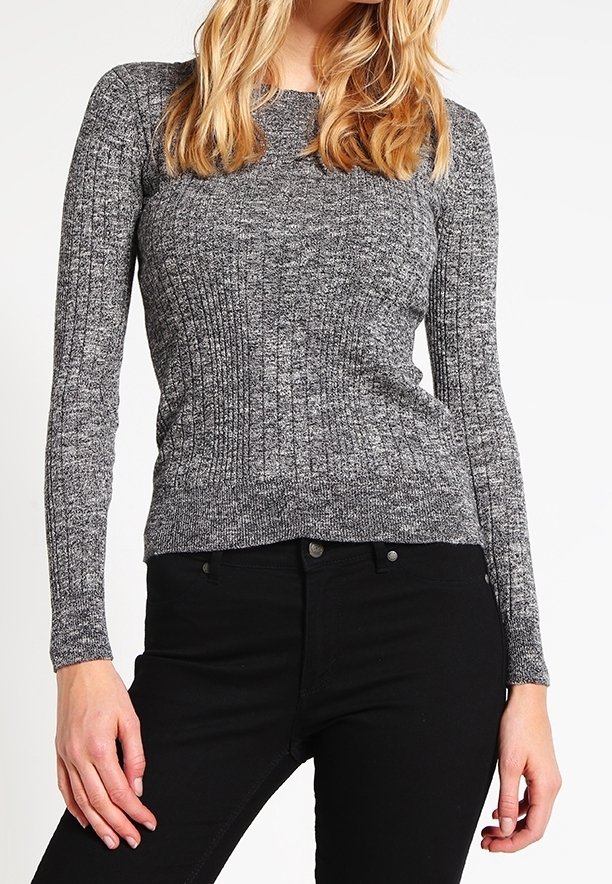 Femme portant un pull gris côtelé ajusté à manches longues et un pantalon noir taille haute, debout les mains relâchées le long du corps.