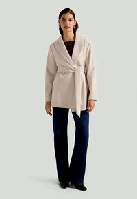Beige wrap coat with long sleeves, belted waist, and notched collar, paired with dark blue flared pants and ankle boots.