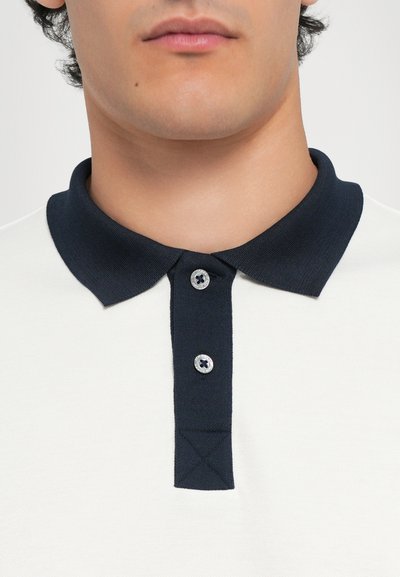 Hvit polo skjorte med marineblå krage, med tre knapper og skrå sømdetaljer. Stoffet ser glatt og litt strukturert ut.
