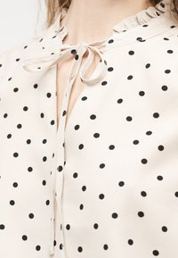 Κρεμ μπλούζα με μαύρες πουά, γιακά με βολάν και κορδόνι στο λαιμό, φορεμένη από άτομο με ορατό λαιμό και σγουρές τούφες μαλλιών.