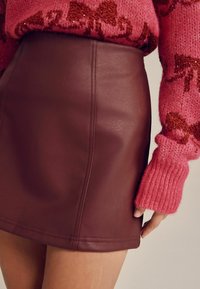 Burgundy läderkjol med en slät yta, försedd med vertikala sömmar. Kombination med en rosa stickad tröja med kontrasterande mönster.