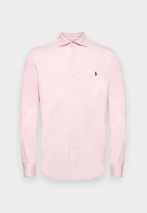 Chemise boutonnière à manches longues rose clair en coton avec une texture douce ; elle possède un col et un petit logo bleu marine sur la poitrine.