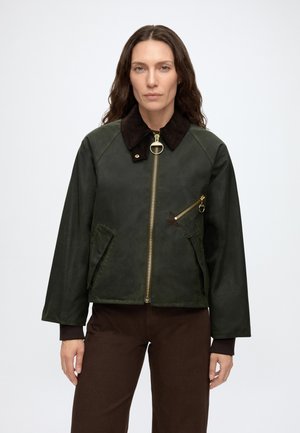 Vrouw met lang bruin haar die een donkergroene jas draagt met een zwarte kraag en gouden ritsen, en een bruine broek, staand tegen een effen achtergrond.