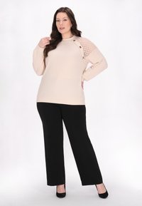 Beige gestrickter Pullover mit strukturiertem Detail an einer Schulter, mit Knöpfen. Kombiniert mit schwarzen weit geschnittenen Hosen und schwarzen Absatzschuhen.