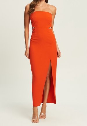 Robe ajustée orange vif sans bretelles avec découpes latérales et une fente haute à l'avant, portée avec des sandales à talons transparents.