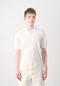 Polo shirt crème à manches courtes avec une patte de boutonnage et une texture douce. Associé à un pantalon blanc cassé, offrant une coupe décontractée.