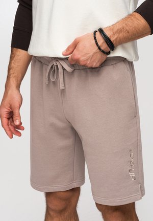 Homme ajustant le cordon de serrage sur un short de sport taupe longueur genoux avec le logo « Level Up » sur la jambe gauche, portant des bracelets noirs en perles et cuir.