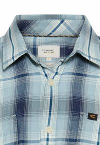 Hellblau kariertes Hemd aus Baumwolle mit weicher Textur. Es hat weiße und marineblaue Streifen, einen knöpfbaren Kragen und eine Brusttasche mit Logo.