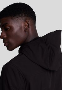 Jachetă neagră cu glugă, cu o textură netedă și un design mulat, cu cusături vizibile pe umeri, fotografiată din lateral.