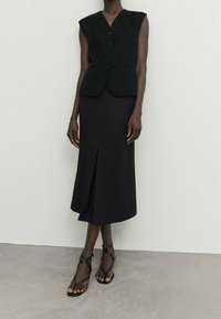 Gilet sans manches noir avec des boutons sur le devant, associé à une jupe noire mi-longue avec une fente sur le côté, accentué par des sandales plates à lanières.
