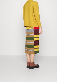 Osoba w musztardowym swetrze, wielokolorowej, paskowanej spódnicy midi i brązowych sznurowanych butach, stojąca na tle jednolitego białego tła.