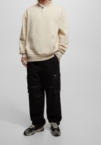 Beige lynlåstrøje med frontlomme, kombineret med sorte cargobukser med flere lommer og hvide syningsdetaljer samt sneakers.