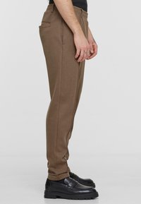 Brown stretch pants with a smooth texture, featuring a side pocket, tapered leg design, and worn with black loafers.