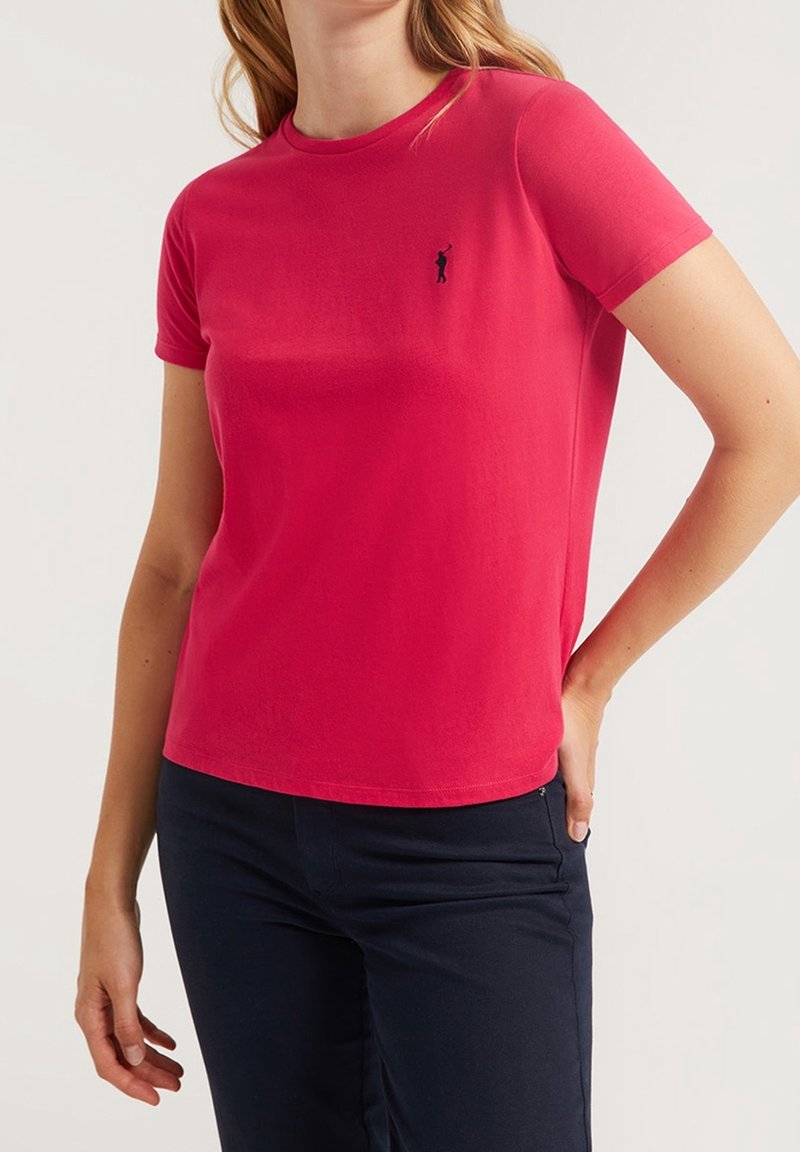 Žena na sobě má jasně červené tričko s krátkým rukávem, na hrudi s malým logem černého pólo hráče, a tmavě modré kalhoty.