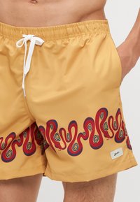 Schwimmshorts in Senfgelb mit einem wirbelnden roten und blauen Schlangenmuster, ausgestattet mit einem elastischen Bund und weißem Kordelzug.