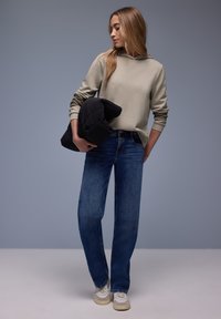 Jeune femme en sweat à capuche beige et jean bleu tenant une doudoune noire, regardant vers le bas sur un fond gris uni.