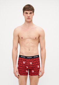 Boxers rouges avec une ceinture noire, présentant un texte répétitif "HUGO" dans différentes tailles et couleurs. Fabriqués en matériau doux et extensible.
