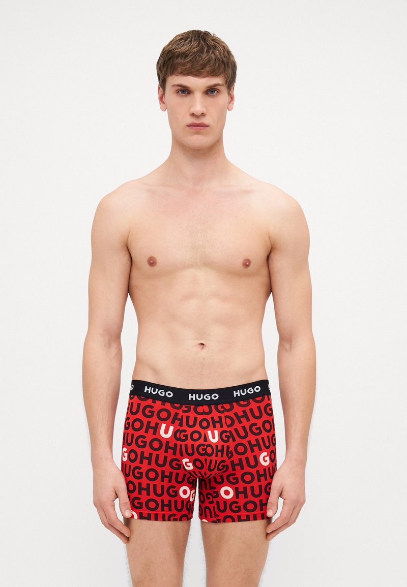 Boxers rouges avec une ceinture noire, présentant un texte répétitif "HUGO" dans différentes tailles et couleurs. Fabriqués en matériau doux et extensible.