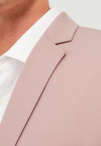 Veste de costume rose clair en tissu texturé, avec un col à revers et des lignes nettes et épurées, portée avec une chemise blanche dessous.