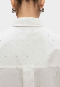 Vrouw van achteren in een wit shirt met kleine metalen studs, zilveren oorhangers en kleinere gouden en zilveren studs in de oorlelletjes.