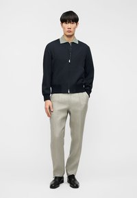 Άνδρας που στέκεται φορώντας ένα σκούρο τζάκετ με φερμουάρ και πράσινο γιακά, ανοιχτό μπεζ παντελόνι και μαύρα επίσημα παπούτσια, σε απλό φόντο.